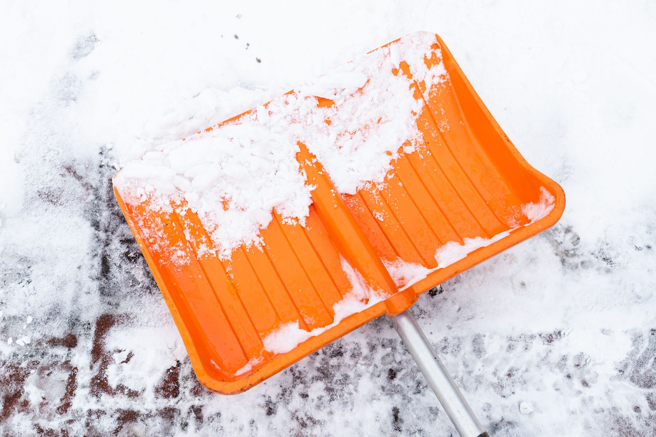 Symbolbild: Orangefarbener Schneeschieber auf einer verschneiten Pflasterfläche